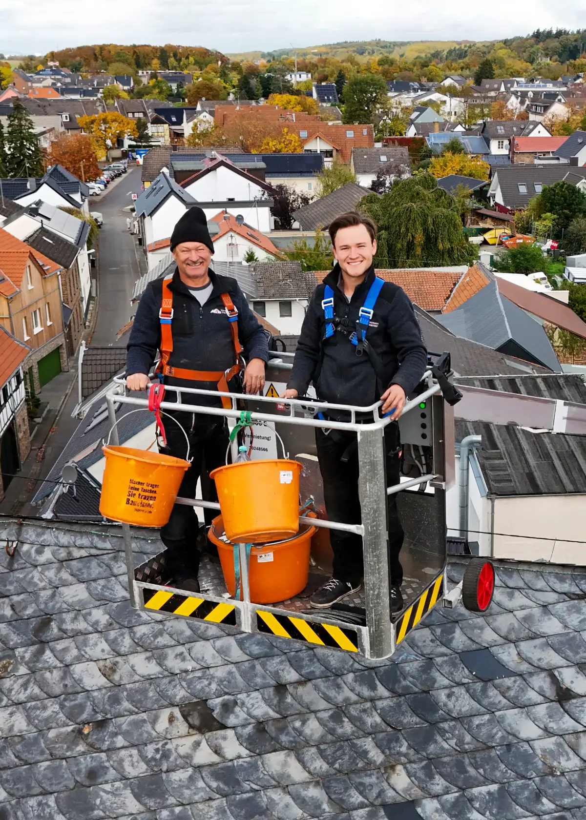 Zwei Personen in Arbeitskleidung mit Sicherheitsgurten stehen mit Eimern im Arbeitskorb über einem Dach, dahinter die Dächer eines Ortes