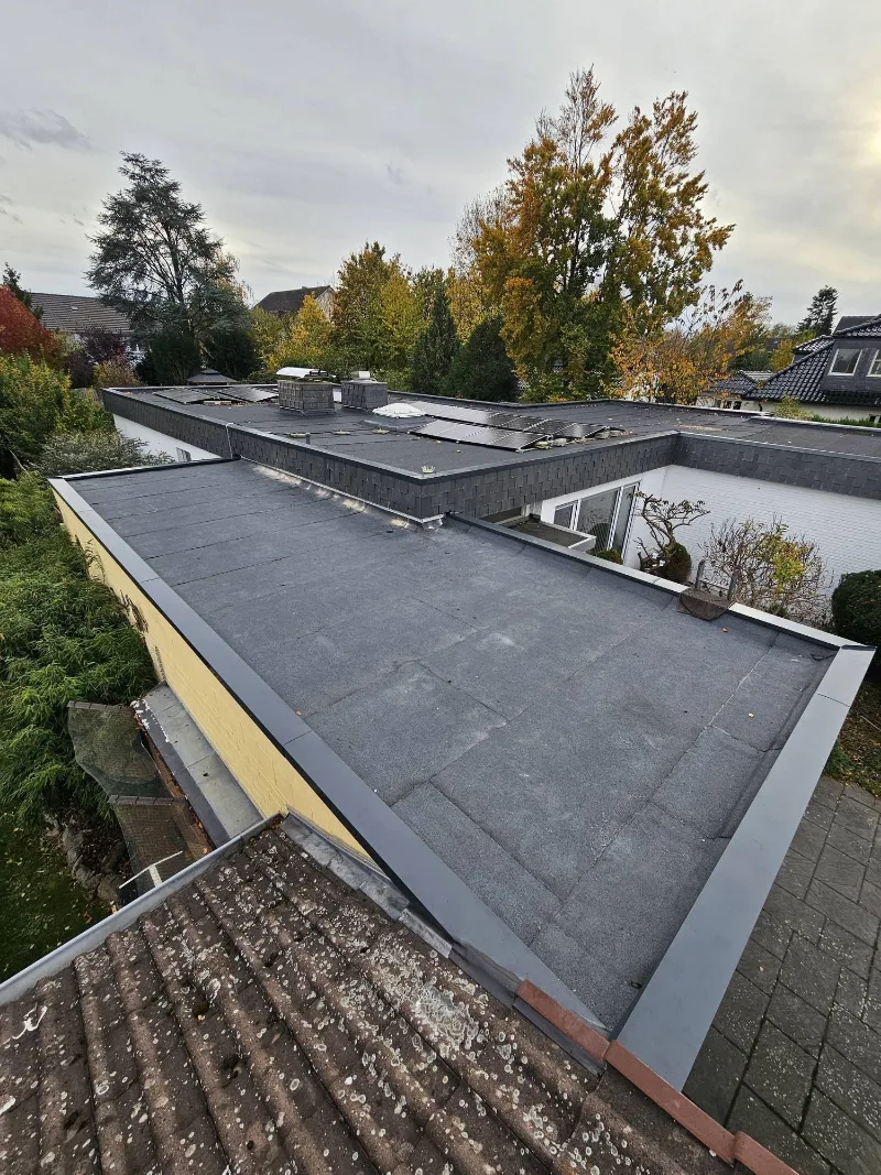 Flachdach mit dunkler Bitumenbahn und Randabschluss aus Metall, im Hintergrund Solarmodule
